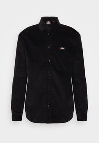 Camicia nera in velluto a coste con bottoni, maniche lunghe, colletto classico e una piccola patch con logo sul petto a sinistra. Tessuto liscio.