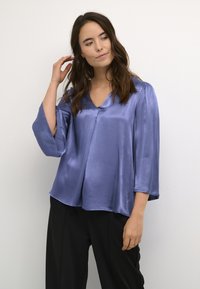 Blusa V- Neck leggera e setosa in lavanda con maniche a tre quarti e orlo leggermente svasato, indossata con pantaloni neri.