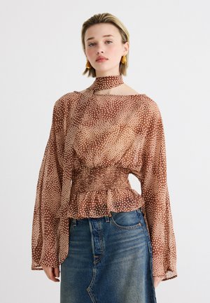 Mladá žena v hnedej blúzke s bodkami a dlhými rukávmi, so zodpovedajúcim šálom na krku, v kombinácii s modrou denimovou sukňou.