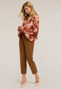 Camicia floreale con dettagli arricciati in rosa e rosso, abbinata a pantaloni marroni a sigaretta e scarpe col tacco nudo.