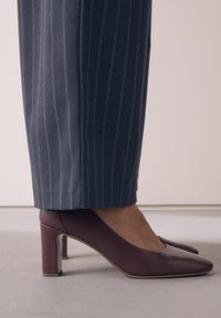Maroon nahka korkokengät teräväkärkisinä ja läpinäkyvällä lohkokorkealla, yhdistettynä tummansinisiin raidoitettuihin housuihin. Suora tekstuuri.