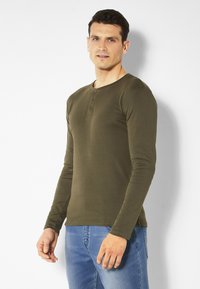 Olivenfarbendes Langarm-Henley-Shirt aus weichem Material, mit Knopfleiste und taillierter Passform. Kombiniert mit blauen Jeans.