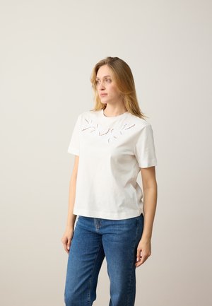 Vrouw met blond haar, gekleed in een wit geborduurd topje met korte mouwen en een blauwe spijkerbroek, staand tegen een effen lichte achtergrond, kijkt naar rechts.