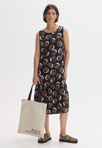 Navy dress with brown and cream circular patterns, gathered waist, sleeveless design. Beige tote bag with black text, worn with brown sandals.