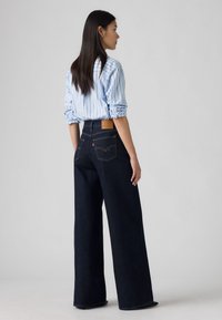 Wijdvallende donkerdenim jeans gecombineerd met een lichtblauwe, gestreepte overhemden met knopen. Kenmerken zijn onder andere opgerolde mouwen en een leren label op de achterkant van de tailleband.