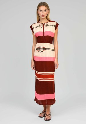 Femme portant une robe sans manches, longue jusqu'aux chevilles, avec des rayures horizontales crème, rose, marron et orange et des motifs géométriques, debout sur un fond gris.