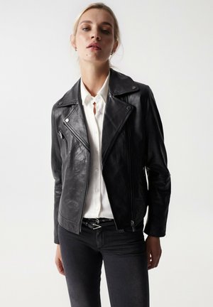 Chaqueta de motorista de cuero negro con cuello de muesca, cremallera asimétrica y bolsillos con cremallera. Usada sobre una camisa blanca y unos jeans oscuros.