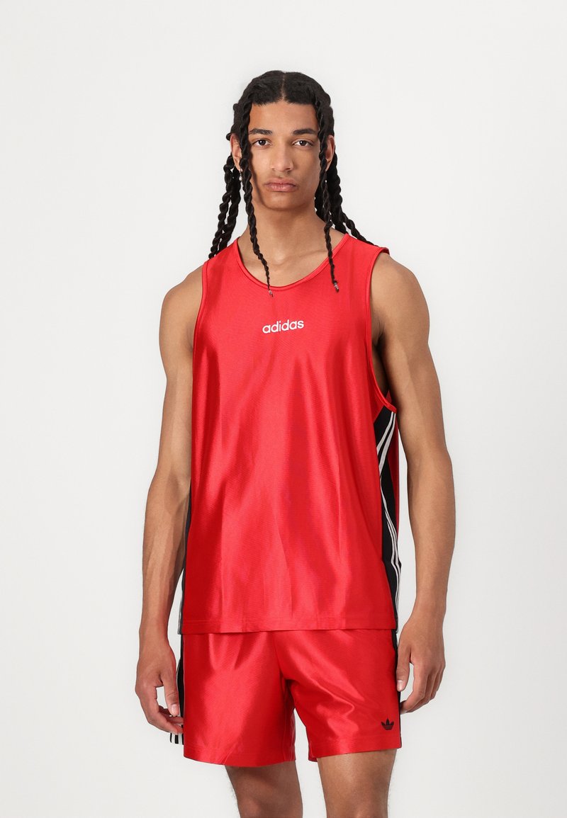 Rdeča športna majica brez rokavov z belim logotipom "adidas" in črnima stranskima črtama. V kompletu z rdečimi kratkimi hlačami, ki sta obe narejeni iz gladkega materiala.