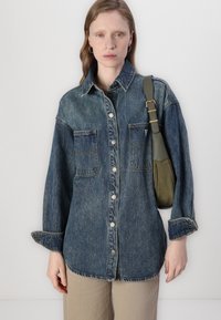 Jeansskjorte i middels blå med knappestolpe, to brystlommer og lange ermer. Kombinert med en grønn skulderveske.