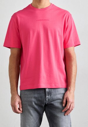 Mężczyzna ubrany w jasnoróżowy t-shirt z krótkim rękawem i subtelnym, umieszczonym na środku logo, w połączeniu z szarymi dżinsami, stoi na tle jasnego, jednolitego tła.