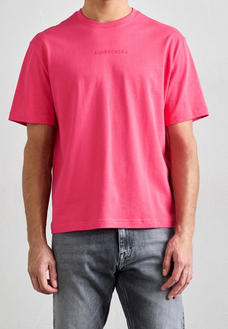 Mężczyzna ubrany w jasnoróżowy t-shirt z krótkim rękawem i subtelnym, umieszczonym na środku logo, w połączeniu z szarymi dżinsami, stoi na tle jasnego, jednolitego tła.