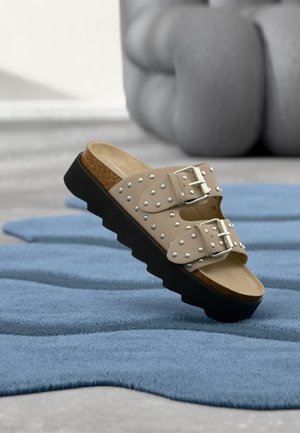 Sandale beige à enfiler avec des clous et des boucles argentés, une semelle intérieure en liège et une semelle extérieure noire rainurée sur un tapis bleu texturé.