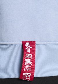 Felpa azzurra chiara con orlo a coste. Presenta un'etichetta in tessuto rossa con scritte bianche che dice "RIMUOVERE PRIMA DEL VOLO". Texture morbida.