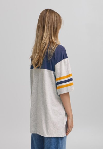 Tricou gri din bumbac cu umeri de culoare albastru marin, având dungi galbene și albastre pe mâneci. Croială lejeră cu o margine rotunjită, purtat cu blugi blue denim.