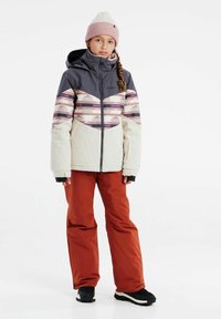 Veste isolée bicolore avec une partie supérieure grise et une partie inférieure crème à motifs ; associée à un pantalon orange et des bottes noires.