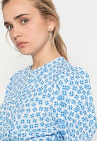 Blusa branca com um padrão floral azul, apresentando um decote redondo, mangas bufantes e tecido texturizado. Brinco de argola prateado visível.