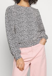 Blusa con stampa leopardata in bianco e nero, con maniche lunghe, scollo rotondo e polsini arricciati. Indossata con pantaloni rosa chiaro.