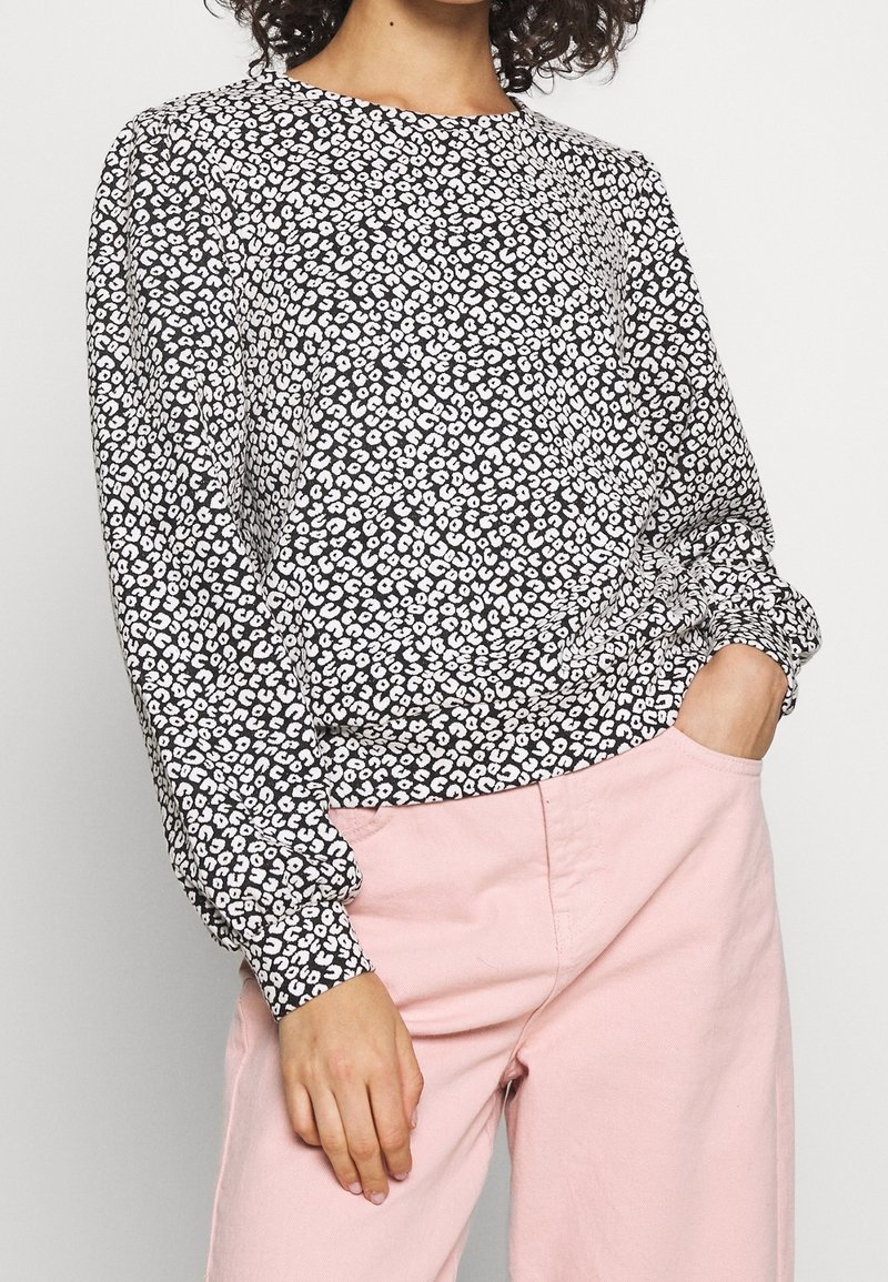 Blusa con stampa leopardata in bianco e nero, con maniche lunghe, scollo rotondo e polsini arricciati. Indossata con pantaloni rosa chiaro.