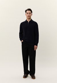 Άνδρας που στέκεται με τα χέρια στις τσέπες, φοράει μαύρο πουλόβερ με φερμουάρ στο γιακά, μαύρο παντελόνι και καφέ επίσημα παπούτσια, μπροστά σε απλό φόντο.