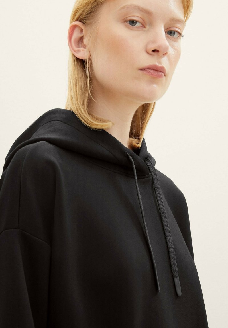 Schwarzer Kapuzenpullover mit lässiger Passform, strukturiertem Stoff und Kordelzügen. Das Design umfasst dezente Nähte an den Schultern.