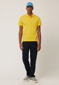 Geel poloshirt met een klein groen logo, gecombineerd met donkerblauwe jeans en beige sneakers. Draagt ook een blauw-wit gestreepte pet.