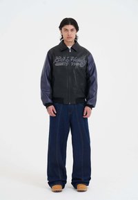 Giubbotto bomber in pelle nera con maniche in pelle viola, caratterizzato da ricamo bianco "Ed Hardy 1977". Abbinato a jeans larghi blu e zoccoli.