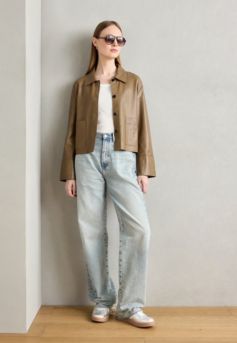 Braune Leder-Cropped-Jacke mit Fronttaschen, kombiniert mit hellblauen, hochgeschnittenen Jeans und metallischen Sneakern. Modell trägt eine Sonnenbrille.