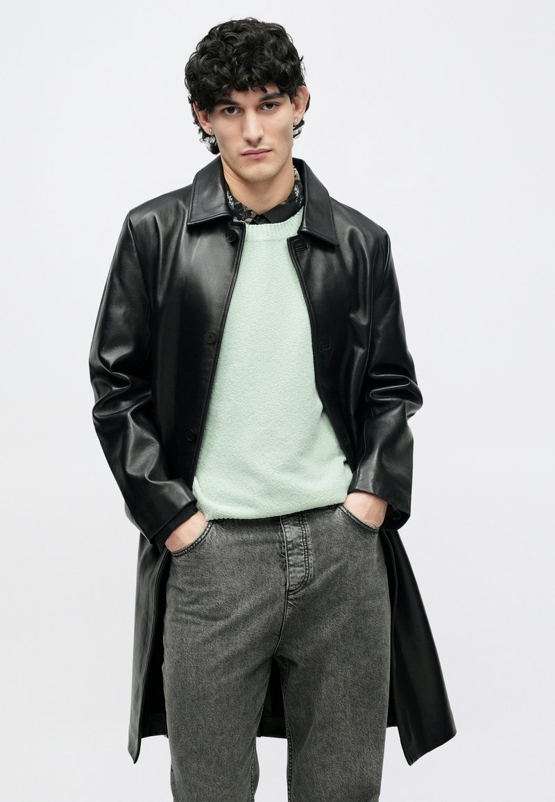 Jeune homme aux cheveux bouclés portant un manteau en cuir noir, un pull vert clair, une chemise noire à motifs et un jean gris, les mains dans les poches.