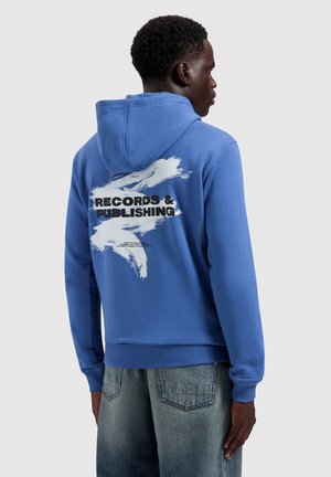 Man in een blauwe hoodie met witte verfstreek en de tekst "RECORDS & PUBLISHING" op de achterkant, gecombineerd met vervaagde blauwe jeans, zijwaarts gericht.