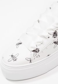 Baskets blanches avec motifs abeilles noirs et larges lacets en satin à volants sur une semelle épaisse et texturée.