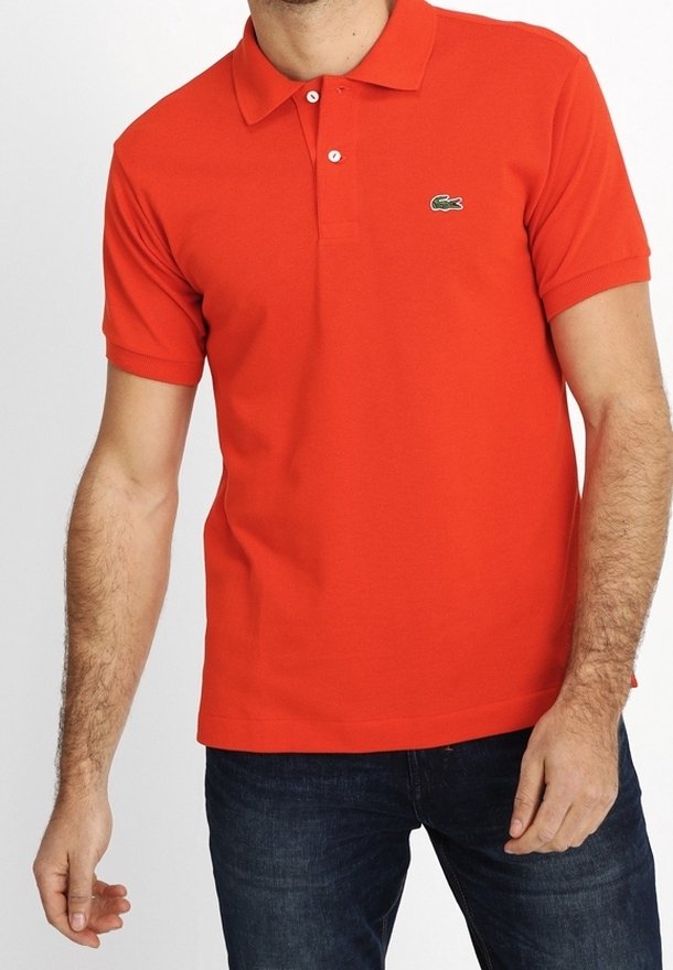 Homme portant un polo à manches courtes orange vif avec un petit logo crocodile vert sur la poitrine gauche et un jean bleu foncé.