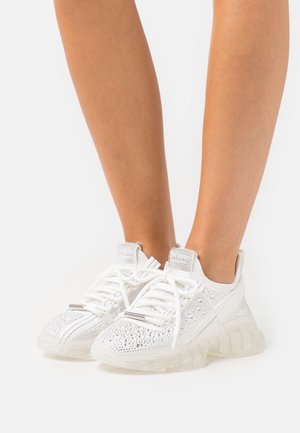 Nogi w białych sneakersach z przezroczystymi, grubymi podeszwami, pokryte srebrnymi cyrkoniami i białymi sznurowadłami, stojące na jednolitym tle.
