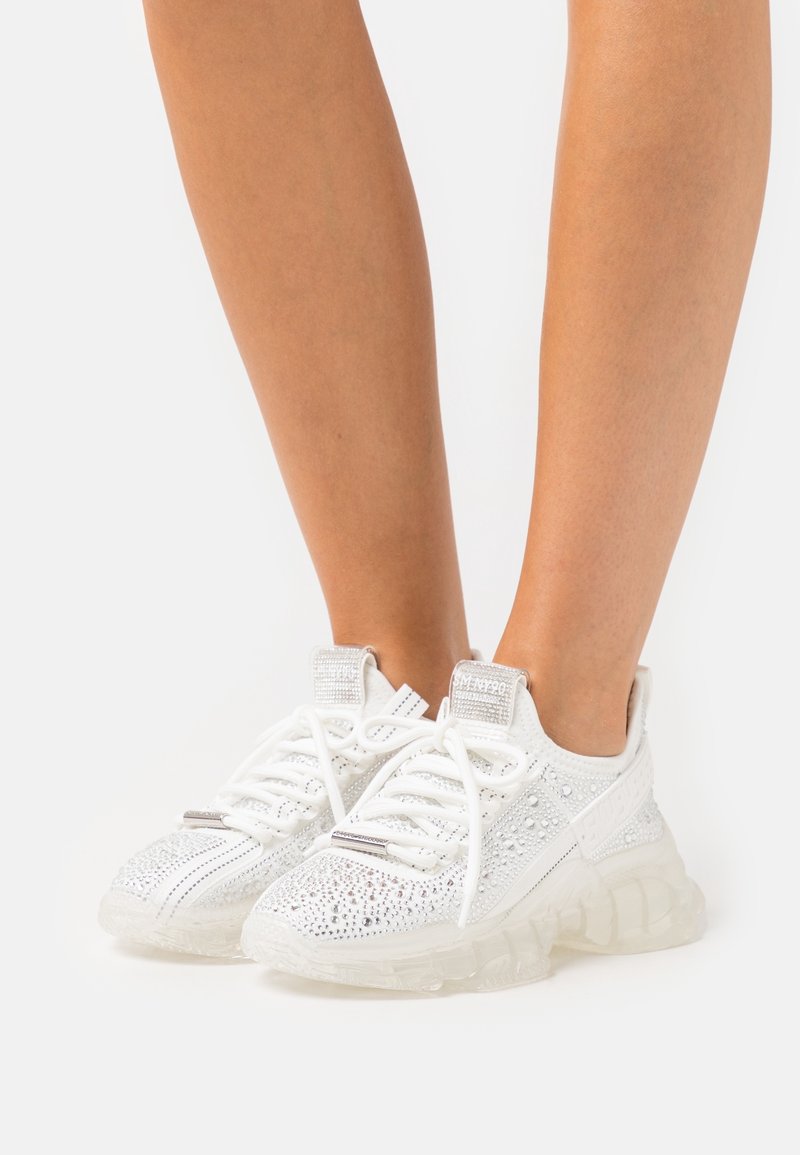 Jambes portant des baskets blanches avec des semelles épaisses transparentes, couvertes de strass argentés et de lacets blancs, debout sur un fond uni.