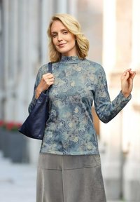 Langarm teal top met bloemenpatroon, opstaand kraagje, zachte textuur, gecombineerd met een grijze rok en een donkerblauwe leren handtas.