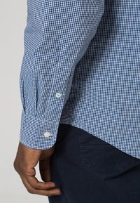 Camisa a cuadros en azul y blanco con puños abotonados, que muestra una tela texturada y botones en contraste. Pantalones oscuros al fondo.