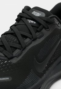 Gros plan d'une chaussure de course noire Nike avec tissu texturé, lacets noirs et logo Nike bien visible sur le côté.
