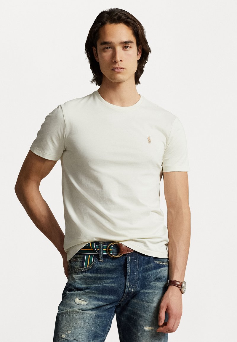 Hvit bomull t-skjorte med rund hals og korte ermer, som har en liten brodert logo på venstre bryst. Brukt sammen med blå slitte jeans.