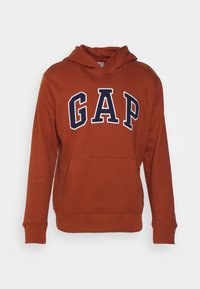 Sudadera de color óxido hecha de tela suave, con un bolsillo canguro y un gran logo "GAP" en azul marino en la parte delantera.