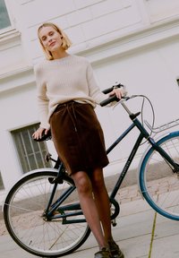 Pull en tricot beige, jupe en velours côtelé marron avec taille à cordon, collants noirs et chaussures. Debout à côté d'un vélo bleu et noir.
