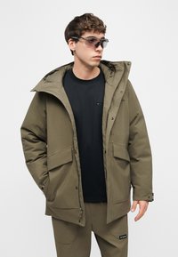 Νέος άνδρας που φοράει παρκά χειμερινό σε ελιά πράσινο, παντελόνι στο ίδιο χρώμα, μαύρο πουκάμισο και σκουρόχρωμα γυ αλιά ηλίου τύπου wraparound, κοιτάζοντας προς τα δεξιά.