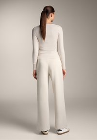 Langärmliges, hellbeiges Rippoberteil kombiniert mit hellen, weiten Hosen. Modell trägt weiße Sneakers; minimalistische Designs und Texturen.