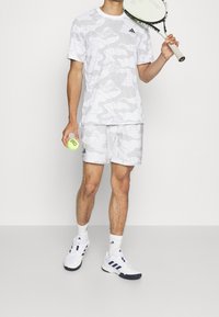 Vitt camouflage-mönstrat träningsplagg, inklusive en t-shirt och shorts. Har en Adidas-logotyp. Vita tennisskor med marinblå accenter, håller en tennisboll.