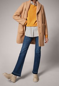 Manteau oversized beige clair, pull orange, chemise à rayures, jeans évasés bleus et bottines à semelle épaisse beige. Tissus texturés et coupe décontractée.