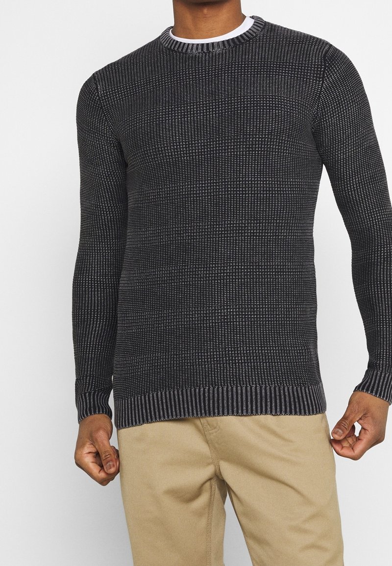 Czarny sweter z dzianiny z fakturowanym wzorem, okrągłym dekoltem oraz ściągaczami przy rękawach i u dołu. Zestawiony z jasnobeżowymi spodniami.