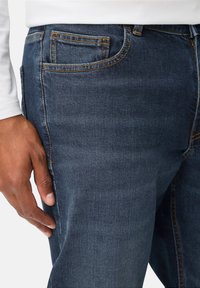Jean en denim bleu foncé avec des coutures jaunes, design à cinq poches, et coupe slim. Vue latérale montrant une texture lisse et un léger dégradé.