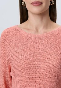 Sweter dzianinowy w miękkim koralowym odcieniu, o luźnym kroju, szerokim dekolcie i teksturowanym wykończeniu. Podkreślony subtelnymi detalami w postaci przeszyć.
