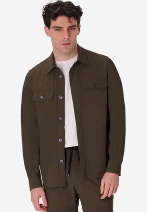 Giovane uomo con capelli corti, ricci e scuri che indossa una camicia verde oliva chiusa con bottoni sopra una t-shirt bianca e pantaloni verde oliva abbinati.