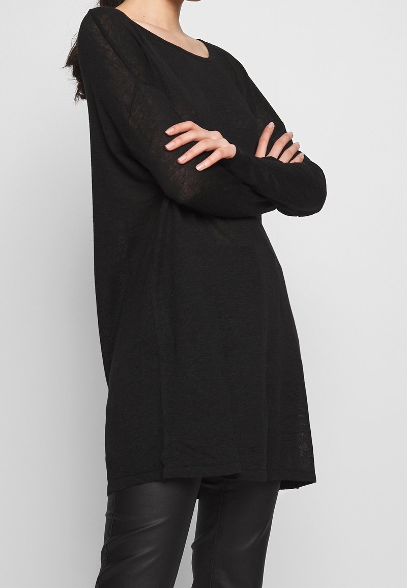Femme les bras croisés portant un long pull noir et un pantalon en cuir noir sur un fond gris clair.