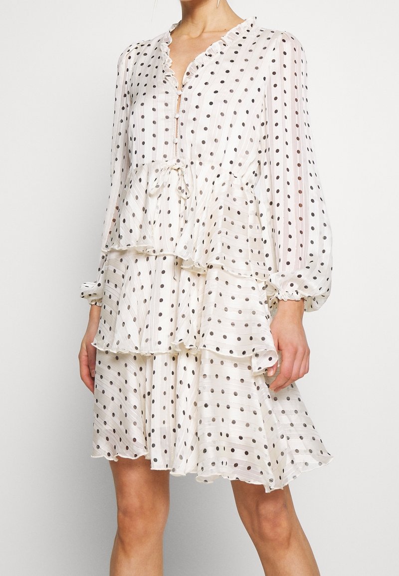 Vestido blanco con lunares negros, con escote fruncido, mangas largas, diseño de volantes y detalle de lazo en la cintura. Hecho de tela ligera.