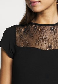 Kvinde iført en sort bluse med blomster-lace yoke og korte ærmer, som viser halsudskæringen samt dele af ansigtet og håret.